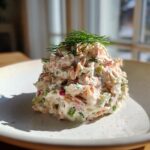 A mound of creamy salmon salad, mixed with celery and herbs, topped with fresh dill, served on a white plate.