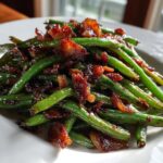 Close-up of bright green beans tossed with caramelized, crispy bacon bits on a white plate.