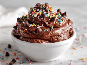 A rich, chocolate brownie batter dip piled high in a white bowl, topped with chocolate chips and colorful sprinkles.