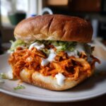 Close-up of a messy, delicious buffalo chicken sandwich piled high with shredded chicken, lettuce, and ranch dressing.