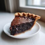 A close-up, appetizing slice of rich chocolate chess pie with a flaky crust resting on a small white saucer.