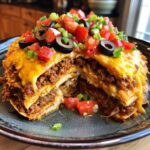 A cross-section view of a layered mexican pizza stack topped with melted cheese, tomatoes, olives, and green onions.