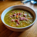 Close-up of a thick, green pea soup topped with diced ham and carrots in a white bowl.