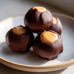 Four chocolate-dipped peanut butter balls stacked on a light plate, showing the creamy peanut butter filling exposed at the top.