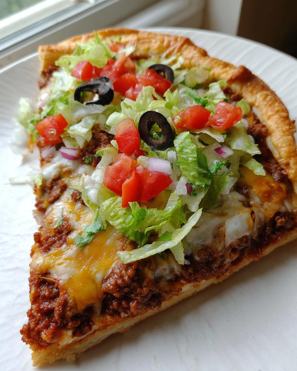 A close-up slice of amazing taco pizza topped with seasoned ground beef, melted cheese, lettuce, tomatoes, and olives.