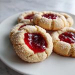 Four delicious thumbprint cookies filled with bright red jam, rolled in coarse sugar, resting on a white plate.