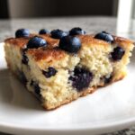 Close-up of a square slice of golden sheet pan pancakes filled with blueberries, served on a white plate.