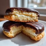 Two halves of a boston cream pie cookies stacked, showing the vanilla cake, cream filling, and chocolate glaze.