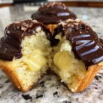 Close-up of a boston cream pie cupcake cut in half showing vanilla cake, yellow pastry cream filling, and chocolate ganache.