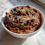 A close-up of rich brownie batter dip topped generously with rainbow sprinkles and chocolate chips in a white bowl.