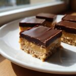 A close-up of a layered buckeye fudge square with a peanut butter base, creamy filling, and chocolate topping.