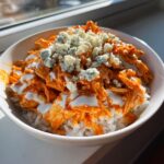 Close-up of a buffalo chicken bowl featuring shredded chicken in orange sauce over white rice, topped with ranch and blue cheese crumbles.