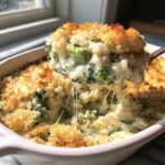 A spoonful of cheesy broccoli rice being lifted from a white baking dish, showing long cheese pulls.