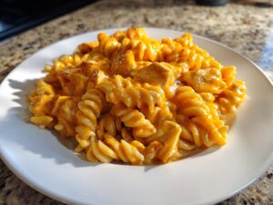 A serving of creamy, orange-sauced cheesy chicken pasta made with rotini noodles on a white plate.