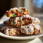 A stack of chewy fruitcake cookies, cut in half to show candied fruit and nuts, dusted heavily with powdered sugar.