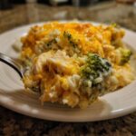 A fork lifting a cheesy bite of chicken broccoli rice casserole, showing melted cheddar cheese and tender chicken pieces.