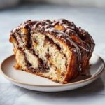 Close-up of a moist slice of chocolate babka showing rich swirls and a sweet white glaze drizzled on top.