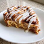 A close-up of a single serving slice of cinnamon roll apple pie topped with thick white icing.