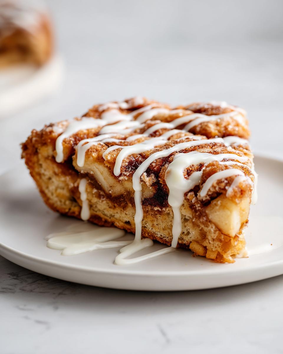 A close-up of a single serving slice of cinnamon roll apple pie drizzled generously with white icing.