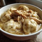 Close-up of a white bowl filled with creamy chicken and dumpling delight, featuring shredded chicken and herbs.