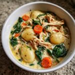 A close-up of a white bowl filled with creamy chicken gnocchi soup, featuring plump gnocchi, shredded chicken, carrots, and spinach.