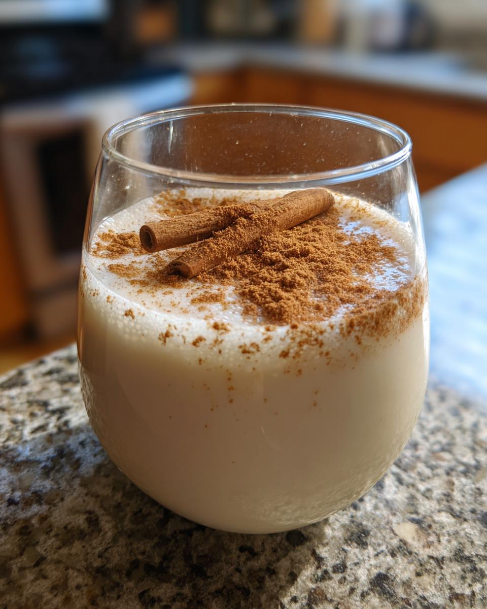 A glass of creamy coquito topped with ground cinnamon and two whole cinnamon sticks.