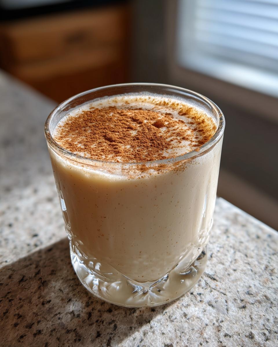 A glass of creamy coquito topped generously with ground cinnamon, sitting on a granite counter.