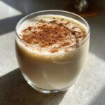 A glass of creamy coquito topped generously with ground cinnamon powder.