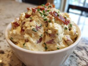 A close-up of rich, creamy million dollar dip served in a white bowl, topped generously with crumbled bacon and fresh green chives.