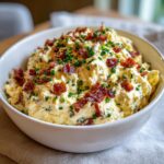 A close-up of creamy million dollar dip generously topped with crispy bacon bits and fresh green chives in a white bowl.