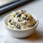 A close-up of a creamy, white olive dip loaded with chopped black olives and green peppers, served in a small white bowl.