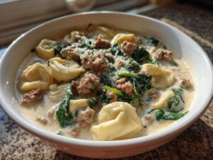 A close-up of a white bowl filled with creamy sausage tortellini soup, featuring tortellini, crumbled sausage, and wilted spinach.