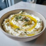 A close-up of creamy whipped feta dip swirled with olive oil and topped with fresh dill and spices.