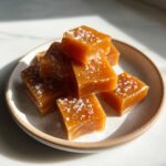 A stack of rich, amber-colored salted caramels sprinkled with sea salt flakes on a small ceramic plate.