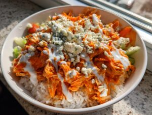 A close-up of a hearty buffalo chicken bowl featuring shredded orange chicken over white rice, topped with blue cheese and ranch dressing.