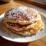 A stack of three golden brown, fluffy pancakes generously dusted with powdered sugar.