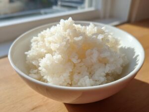 Close-up of perfectly cooked, fluffy sushi rice mounded high in a small white bowl, illuminated by sunlight.