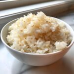 A close-up of a white bowl filled with fluffy, perfectly cooked sushi rice sprinkled with seasoning.