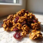 A close-up pile of crunchy, golden-brown gingerbread granola clusters mixed with dried cranberries.