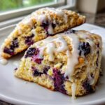 Two triangular blueberry scones drizzled heavily with white vanilla glaze, served on a white plate.
