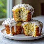 Three delicious lemon poppy seed muffins stacked on a white plate, topped with thick white glaze and visible poppy seeds.