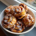 A close-up of several gooey cinnamon roll bites drizzled with white icing, served in a small white bowl.