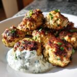 A close-up of several golden-brown Greek chicken meatballs served on a white plate with a dollop of herbed yogurt sauce.