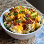 A close-up of loaded baked potato salad topped with shredded cheddar cheese, bacon bits, and green onions.