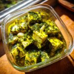 Close-up of cubed marinated cheese soaking in bright green herb oil with whole garlic cloves.