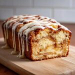 A loaf of freshly baked apple fritter bread, sliced to show apple filling, drizzled with white glaze.
