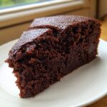 A close-up of a rich, dark slice of moist chocolate cake recipe on a white plate.