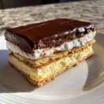 A layered slice of no-bake eclair cake featuring graham crackers, vanilla cream, whipped topping, and rich chocolate glaze.