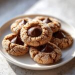 A close-up of several sugary peanut butter blossoms cookies topped with melted chocolate kisses on a white plate.