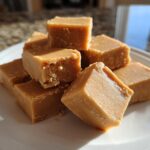 A close-up stack of creamy, square peanut butter fudge bites resting on a white plate.